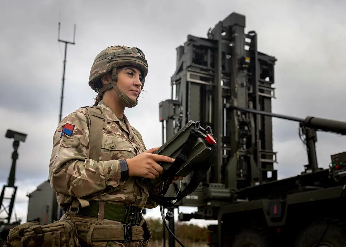 A female soldier from the Royal Artillery, wearing a helmet and camouflage uniform, holds a control panel for the Sky Sabre air defence missile system. The system is mounted on a truck in the background.
