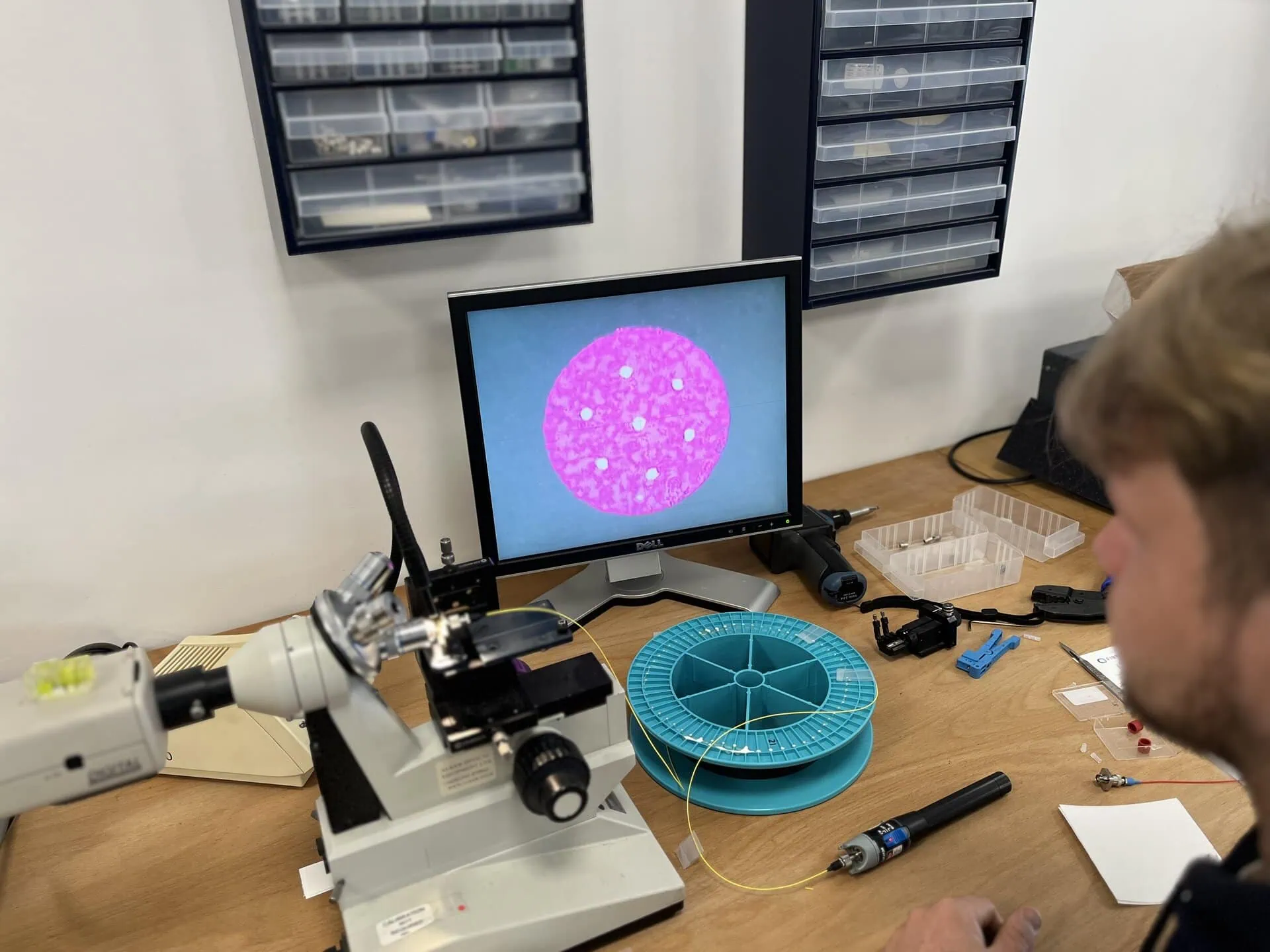 A technician sits at a workbench looking at a computer monitor that displays a magnified view of a fibre optic end-face. On the desk are a microscope, a light blue reel of yellow fibre optic cable, and various tools.