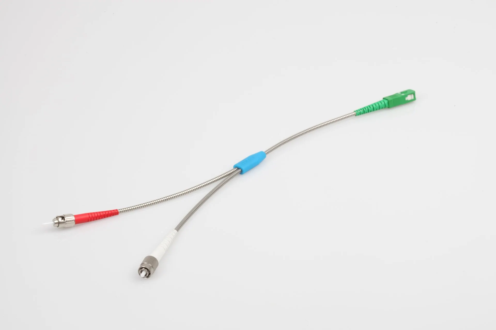 A detailed product shot of a fibre optic Y-splitter cable. A single metal-braided cable with a green SC/APC connector splits into two thinner cables, terminating in a red connector and a white/silver FC connector.