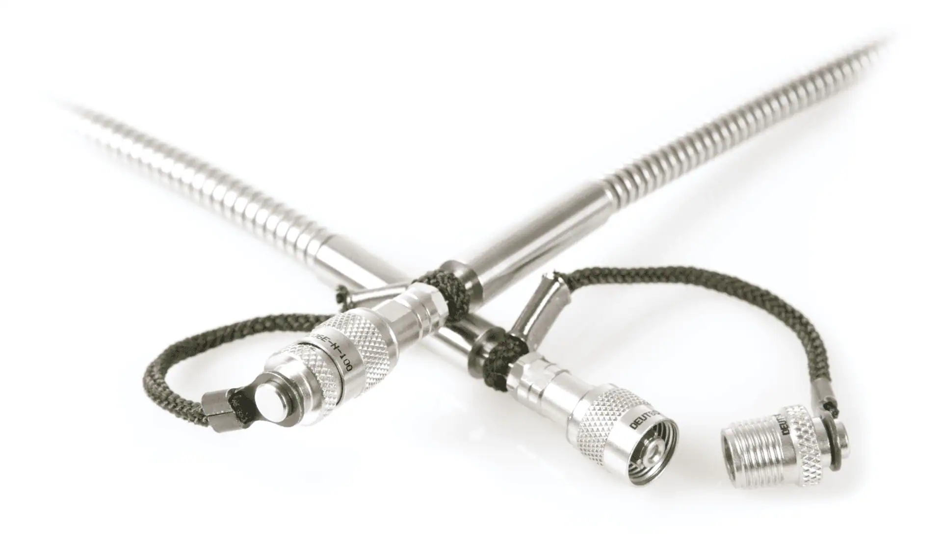 A close-up shot of two braided metal cables crossing, each ending in a specialized ruggedized metal connector. A green cord is tied around the junction, and a separate connector cap is shown next to the right-hand cable.