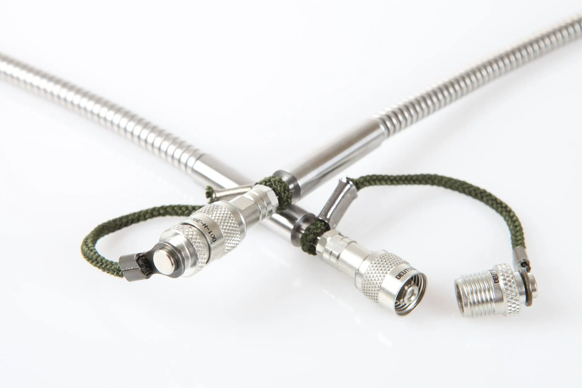 A close-up shot of two braided metal cables crossing, each ending in a specialized ruggedized metal connector. A green cord is tied around the junction, and a separate connector cap is shown next to the right-hand cable.