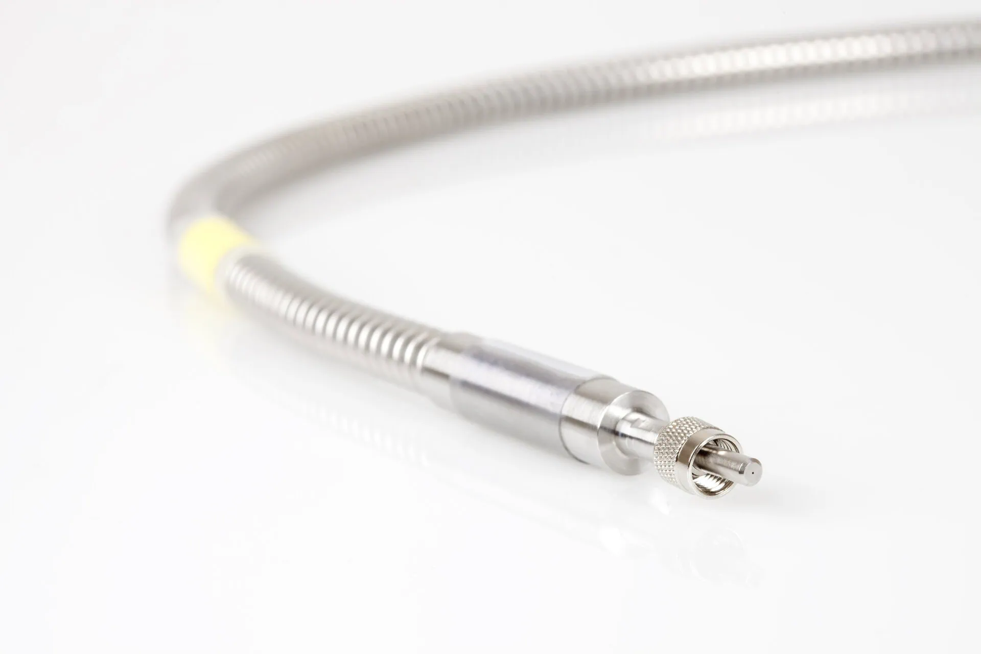 A detailed shot of a coiled, braided metal fibre optic cable that terminates in a cylindrical metal connector with a small ferrule visible at the end. A yellow label is on the cable's sheath.
