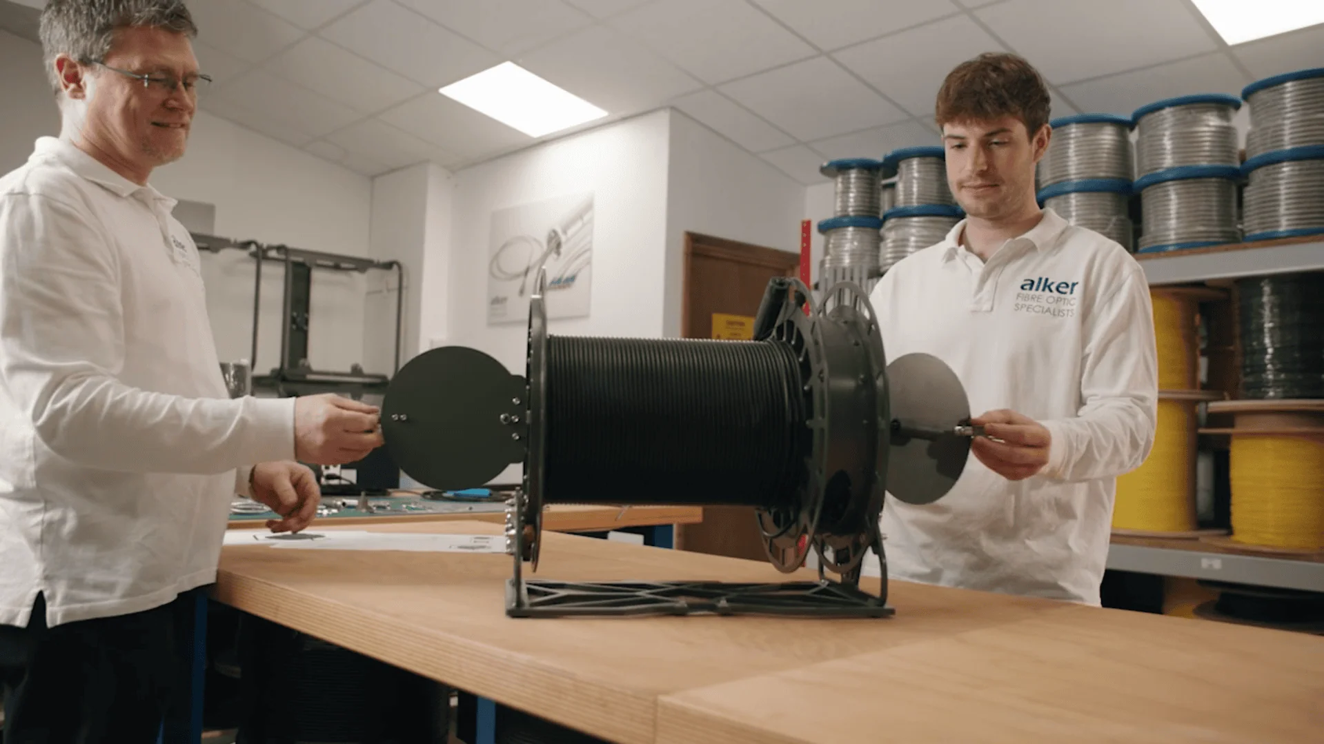 Two male Alker technicians, wearing white company polo shirts, are shown working together to assemble a large, black cable reel on a workbench in a workshop filled with various coils of fibre.