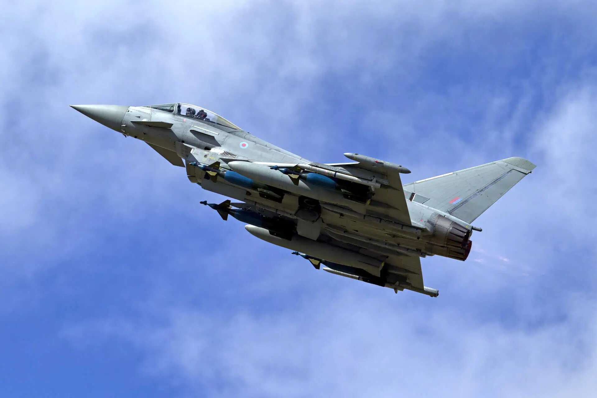An RAF Eurofighter Typhoon fighter jet, armed with missiles, is shown flying against a blue sky with scattered clouds.