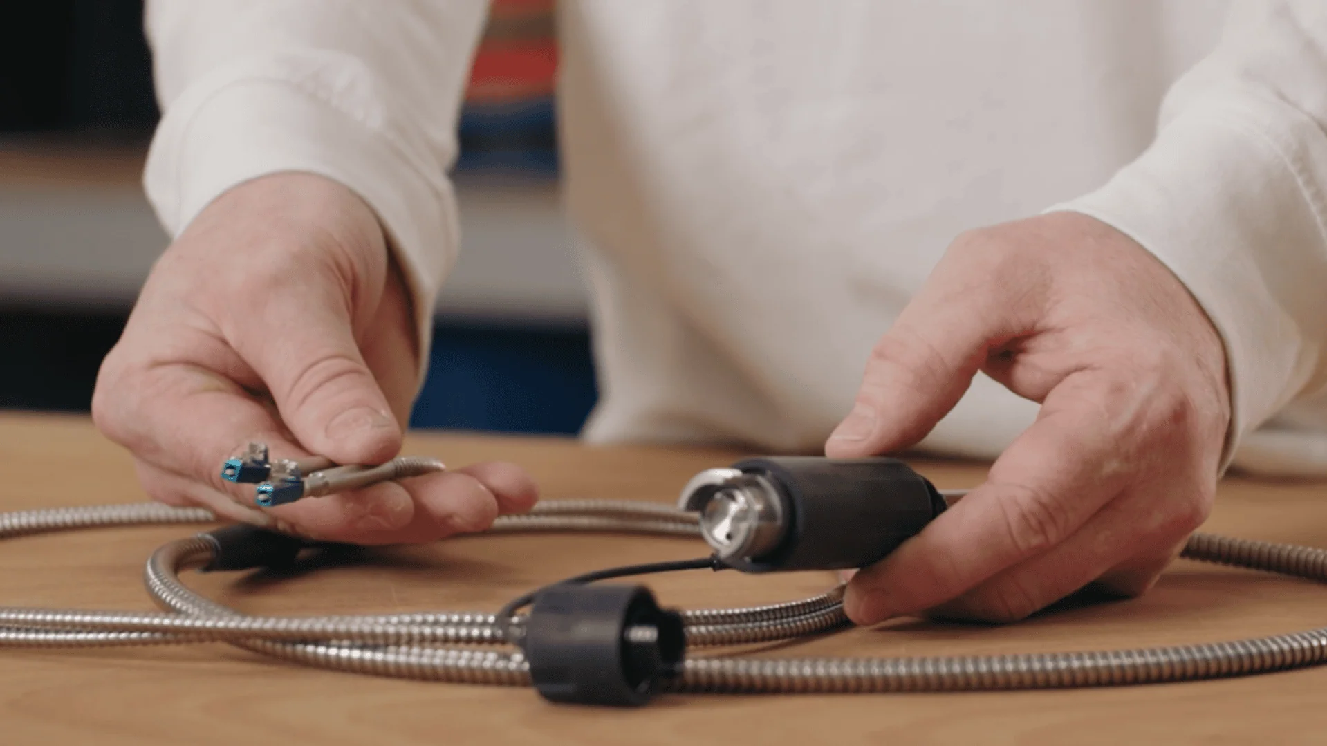 A close-up of a person's hands connecting a ruggedized fibre optic cable. The left hand holds a pair of blue LC connectors, while the right hand holds a black cylindrical connector body attached to a coiled, metal-braided cable.