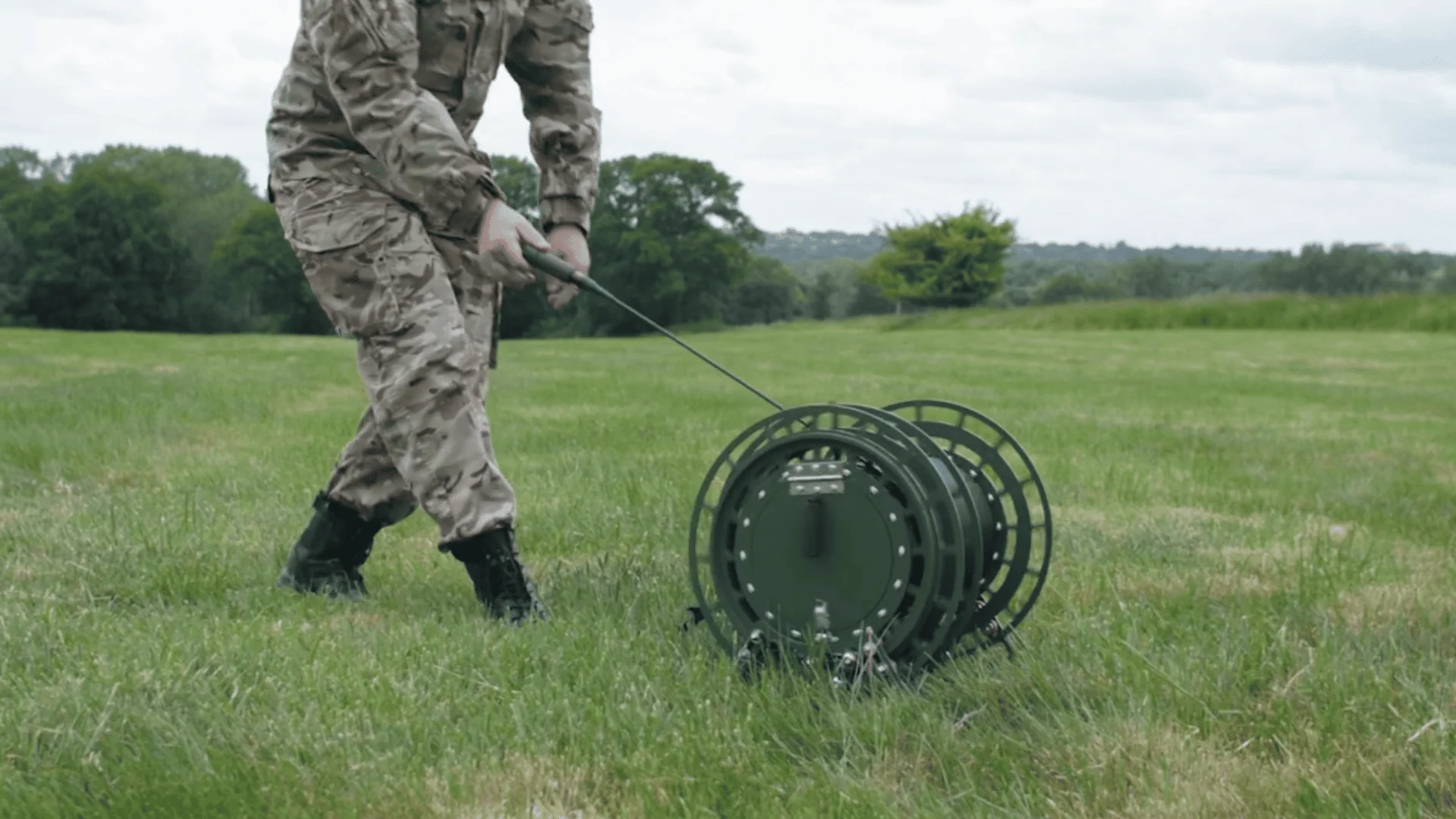 A soldier in a camouflage uniform is shown from the knees down, pulling a large, wheeled, olive-green fibre optic cable dispenser across a grassy field.