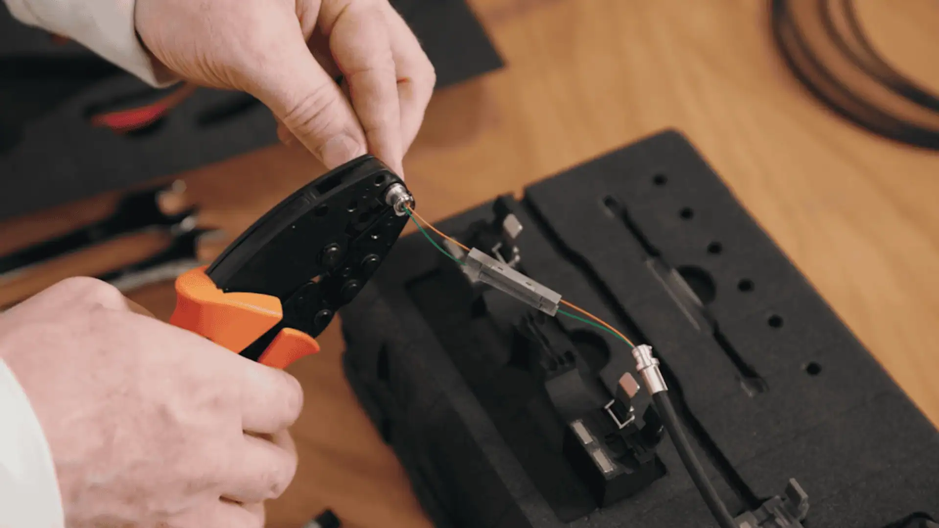 A close-up shot of a technician's hands using a bright orange crimping tool to secure a connector onto a multi-strand fibre optic cable held in a specialized jig.
