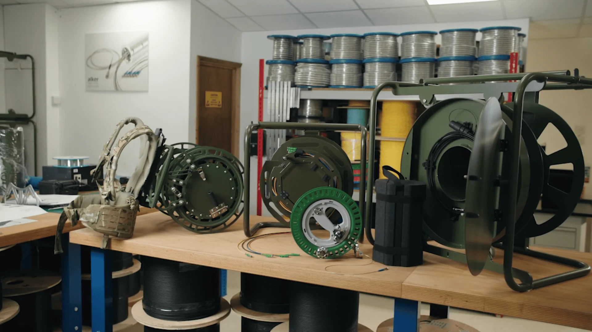 A collection of military and field-grade fibre optic equipment is arranged on a workbench, including several tactical cable dispensers of varying sizes=