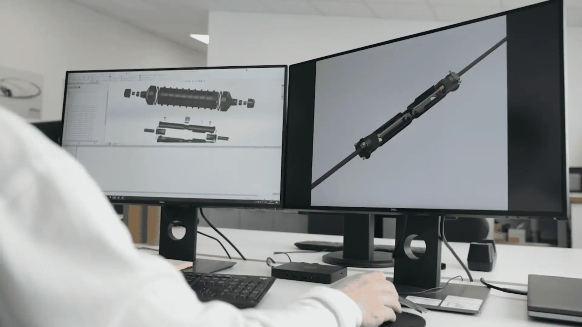 A person's hands are shown using a mouse and keyboard at a dual-monitor computer setup. Both screens display a 3D CAD model of a complex mechanical component, illustrating engineering design and product development.