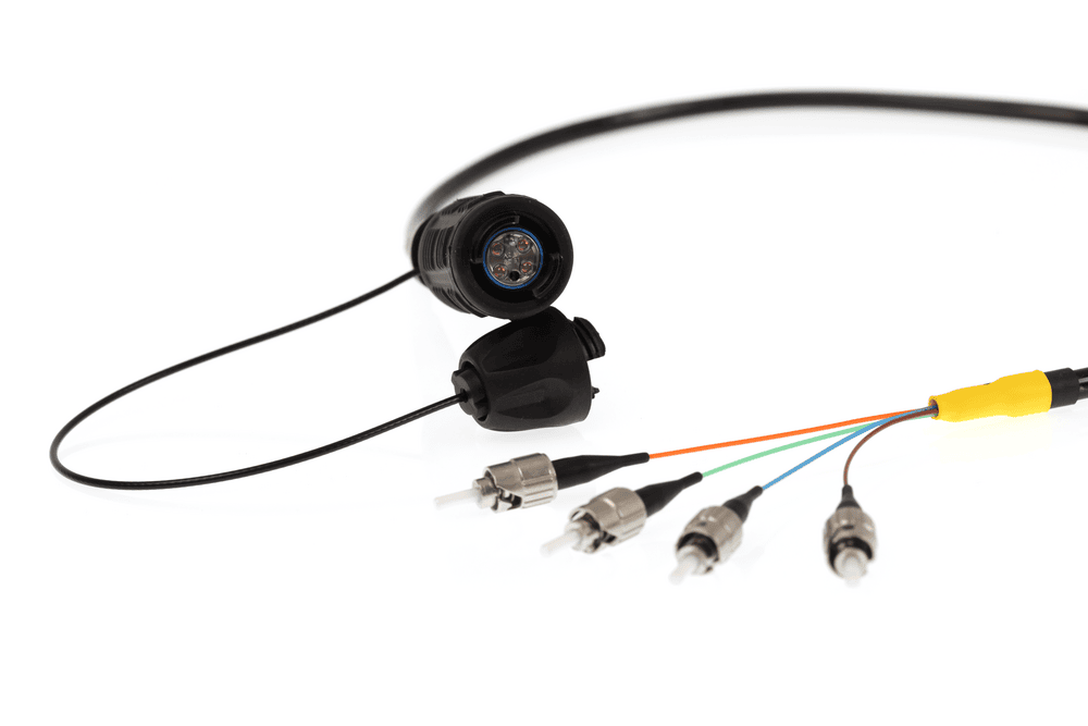 A specialized fibre optic breakout cable is shown with a thick black main cable ending in a ruggedized connector. Four individual colored fibres (yellow, orange, green, and blue) emerge from the connector, each terminating in a separate metal FC/ST style connector.